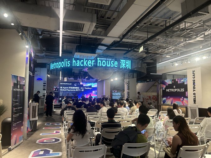 An audience seated in white chairs facing a stage with a digital screen at the 'Metropolis hacker house 深圳' event in Shenzhen, featuring a 'Hello Young Cafe' area to the right.