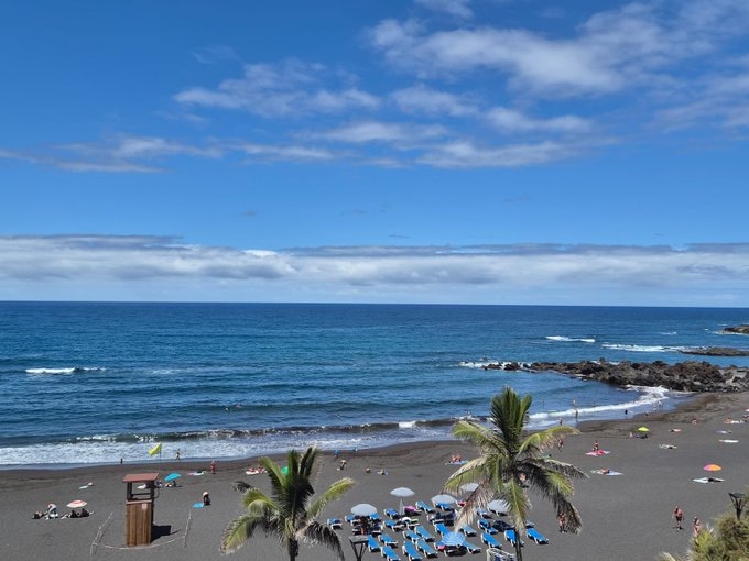 A scenic view of a beach with dark sand, palm trees, lounge chairs, and umbrellas, under a partly cloudy blue sky.