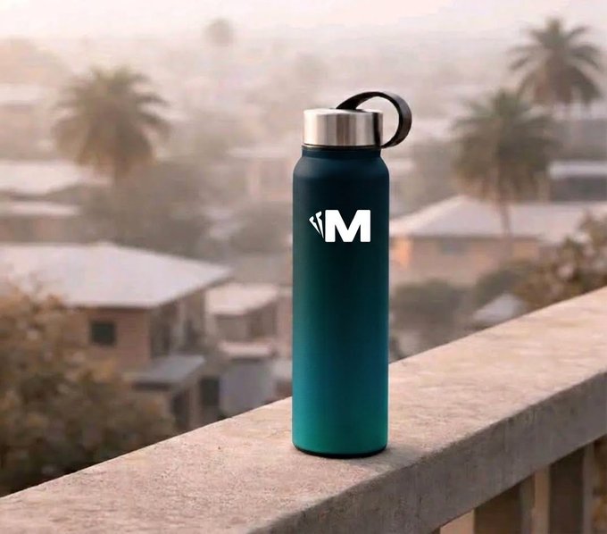 A teal gradient insulated water bottle with a silver cap and a stylized 'M' logo sits on a concrete ledge overlooking a hazy cityscape with palm trees.
