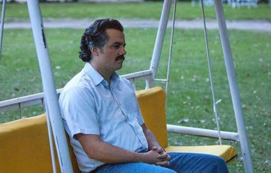 A man with a mustache sitting on a yellow swing bench in a park, looking off to the side in a contemplative pose.