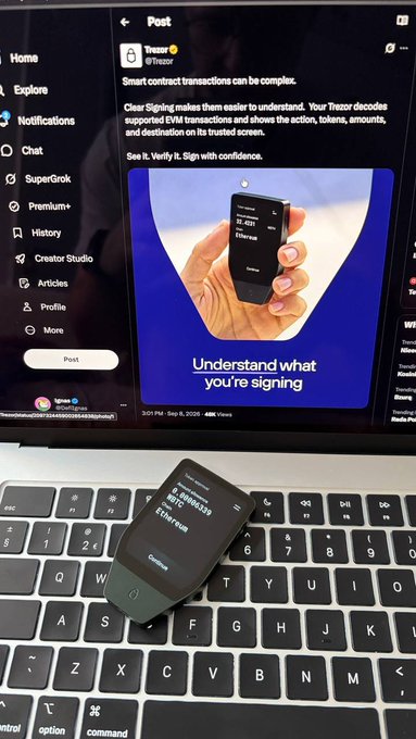 A black Ledger hardware wallet displaying a transaction for "0.000369 ETH" rests on a MacBook keyboard, while the screen behind shows a Trezor post titled "Understand what you're signing," emphasizing the importance of verifying transactions rather than blindly signing them to avoid malicious exploits.