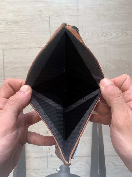 Two hands hold open a brown leather wallet or cardholder, revealing a black, geometric, diamond-shaped design featuring a Cardano (ADA) logo etched in the center.