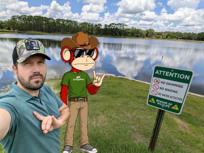 A man in a teal polo and camo cap takes a selfie beside a cartoon monkey mascot wearing a cowboy hat and "JungleBay" shirt, posing by a swampy pond with a sign reading "ATTENTION: NO SWIMMING, NO WADING, NO WATER ACTIVITIES, BEWARE OF WILDLIFE." The scene humorously visualizes the post's mention of a "swamp" setting, tying into the JungleBayAC web3 PFP branding.