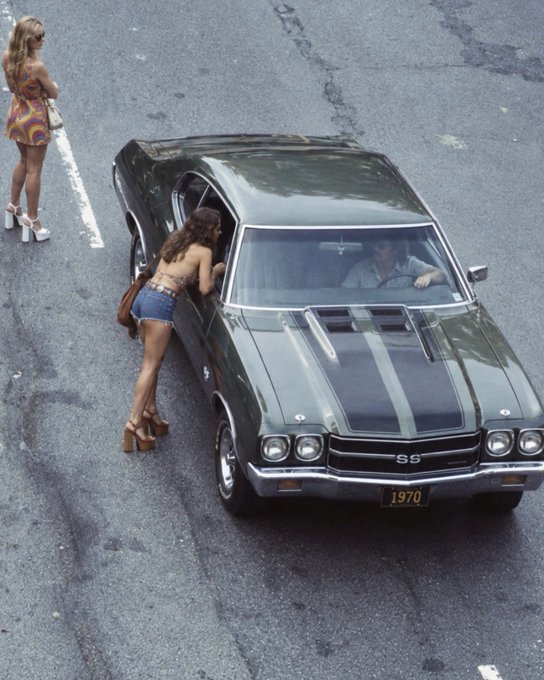 A vintage-style overhead photo of a 1970 Chevrolet Chevelle SS muscle car with two women in 1970s fashion standing beside it.