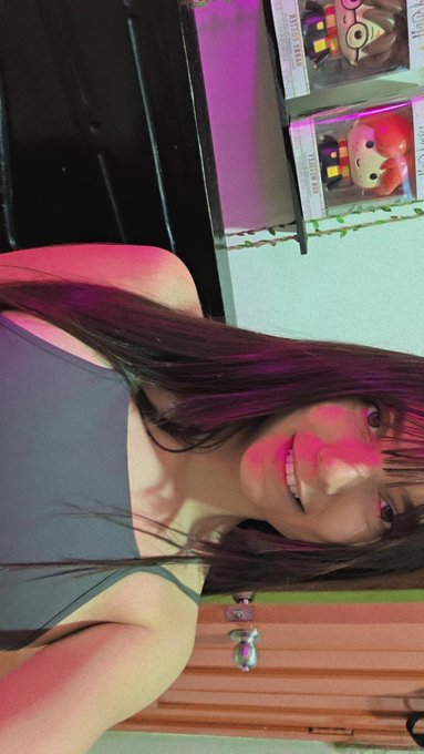 A selfie-style photo of a young woman with long dark hair in a gray top, with shelves of merchandise visible behind her in pink lighting.