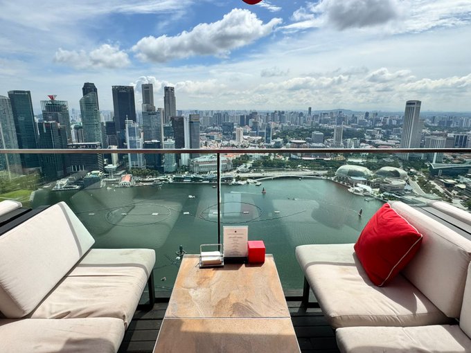 A balcony view overlooking a city skyline and a body of water, with outdoor seating and a table in the foreground.
