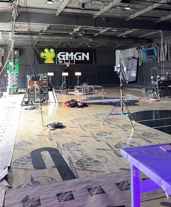 Behind-the-scenes look at a GMGN studio space with filming equipment, lighting rigs, and a basketball court floor featuring a 2K20 logo and tournament branding.