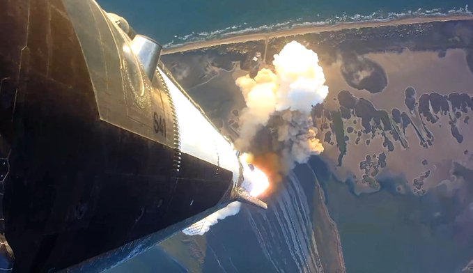 An aerial or onboard view from a spacecraft, likely a SpaceX rocket, showing an engine firing with a plume of fire and smoke against a backdrop of land and water below.