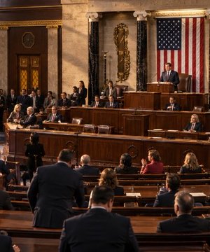 The image shows the U.S. House of Representatives chamber with members gathered, an American flag displayed prominently, and a speaker addressing the assembly. This setting relates to the breaking news about two new crypto bills advancing through the House, illustrating the legislative environment where these cryptocurrency regulations are being deliberated and voted upon.