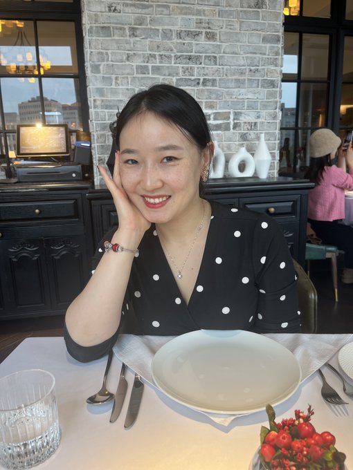 Image 6: A portrait of a smiling Asian woman wearing a black dress with white polka dots, sitting at a restaurant table set with an empty plate and silverware. She has red lipstick, a delicate necklace, and rests her hand near her face.