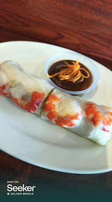 Two fresh summer rolls filled with shrimp and vegetables are served on a white plate alongside a small bowl of dark soy-based dipping sauce, captured on a Seeker/Solana mobile device.