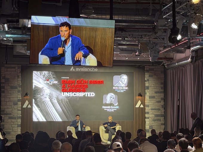 The image shows Emin Gün Sirer, CEO of Ava Labs, speaking on stage at a New York event titled "Emin Gün Sirer & Haseeb Qureshi Unscripted," with his quote about targeting the enterprise side and unified ecosystems displayed on the screen behind him.