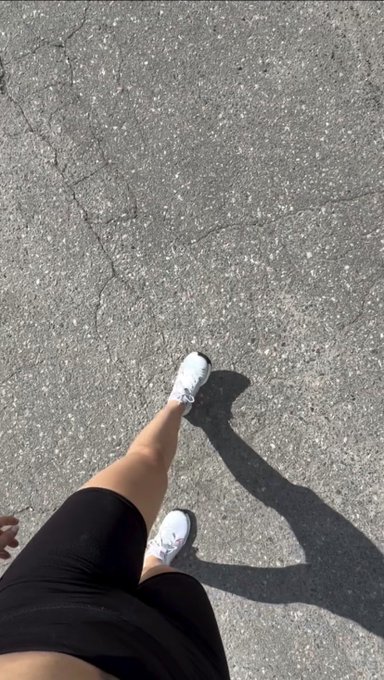 A first-person view looking down at legs in black shorts and white sneakers walking on cracked asphalt, with the person's shadow cast on the ground.