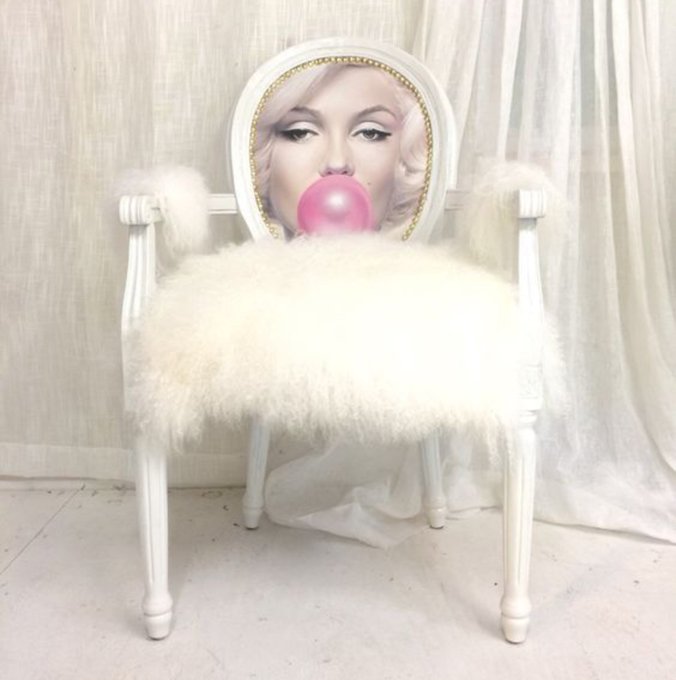 An ornate white chair with a fluffy white seat cover, featuring a painted portrait of Marilyn Manson blowing a pink bubblegum bubble on the backrest, set against sheer white curtains.