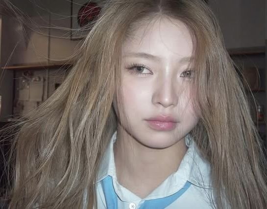 A close-up selfie of a young woman with long, messy blonde-brown hair, wearing heavy eyeliner and pink lipstick, dressed in a light blue collared shirt.