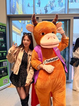 A young woman poses making a peace sign next to a person in a brown cow mascot costume wearing a 'TurboTax' branded sash at what appears to be an indoor event.