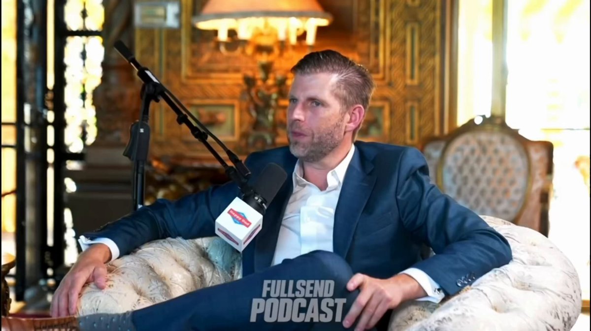 The image features a man sitting in a chair, wearing a suit and tie. He has his legs crossed while looking off into the distance with an expression of thoughtfulness or contemplation. The background includes a couch and a lamp, suggesting that he is in a living room setting. The image also contains text that reads "FullServe Podcast" and "ERIC TRUMP SAID: You COULD SEND ME A MILLION DOLLARS IN $BTC RN AND IT’D HIT MY WALLET IN 3 SECONDS, AND YOU PRACTICALLY PAY ZERO FEES DURING THE PANDEMIC. Focus on what is in the image and how it relates to the post."