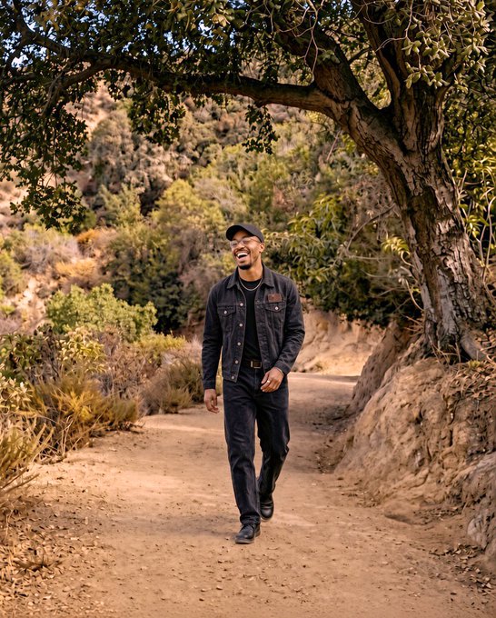 A smiling man wearing a black cap and dark denim jacket walks along a dirt path under a large tree, set against a backdrop of a wooded hillside.
