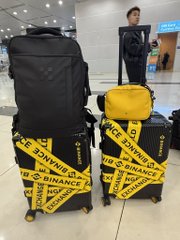 A person stands behind two pieces of luggage wrapped in yellow tape featuring the 'BINANCE' logo in an airport terminal.