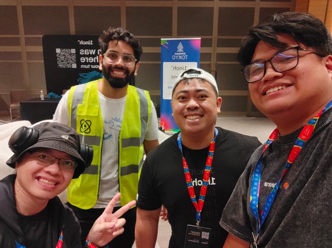 Four smiling men pose together at what appears to be a tech event, with one wearing a yellow safety vest with a React logo, and Tokyo brand banners visible in the background.