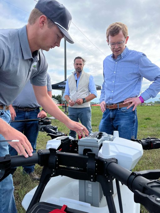 Three men examine a large agricultural drone outdoors, with one person pointing at the drone's internal components while the others observe closely.