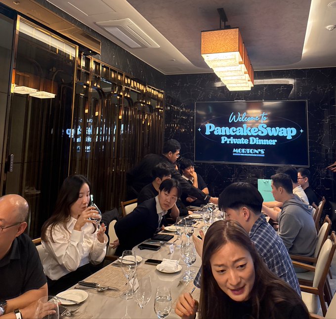 A lively dinner scene at Morton's Steakhouse where attendees are seated around a long table set with wine glasses and plates, with a screen in the background displaying "Welcome to PancakeSwap Private Dinner - MORTON'S," marking the DEX's first private dinner gathering.