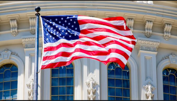 The image features the American flag waving in front of what appears to be the U.S. Capitol building, symbolizing the legislative and governmental backdrop against which the CLARITY Act debate unfolds. This visual reinforces the post's focus on Senate leaders Thune and Schumer, highlighting the political arena where the National Sheriffs' Association is voicing concerns over Section 604's potential exemptions from KYC, AML, and registration requirements in the crypto industry.