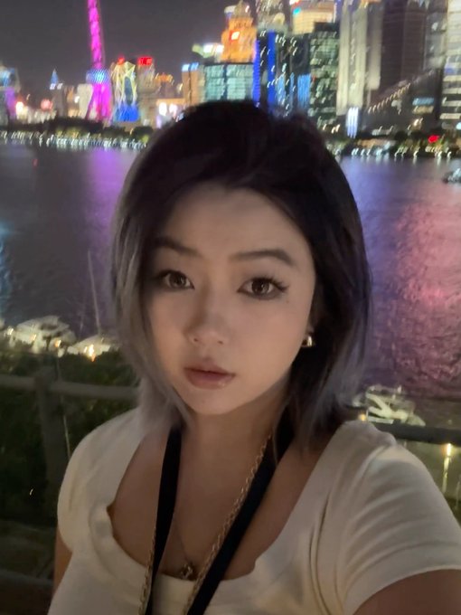 A selfie of a young woman with two-toned hair posing in front of a brightly lit city skyline at night, likely the Shanghai Bund.
