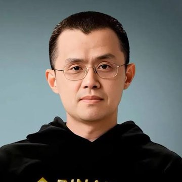 A portrait photograph of an East Asian man with short black hair wearing glasses and a black hoodie with partially visible 'BINANCE' logo, photographed against a gray background.