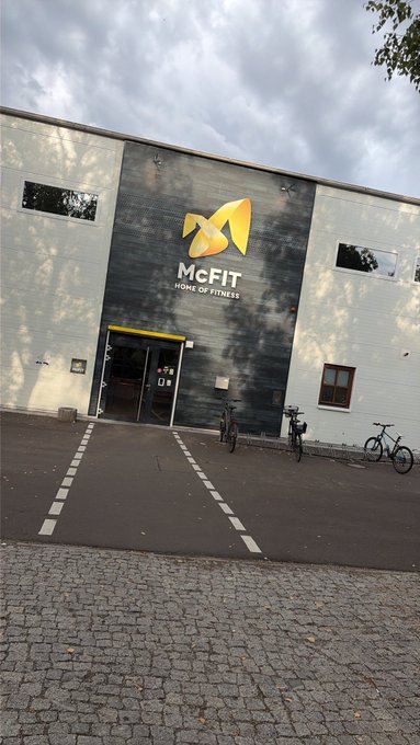 Exterior view of a McFIT 'Home of Fitness' gym building with dark wood-paneled facade, featuring the yellow McFIT logo and bicycles parked outside.