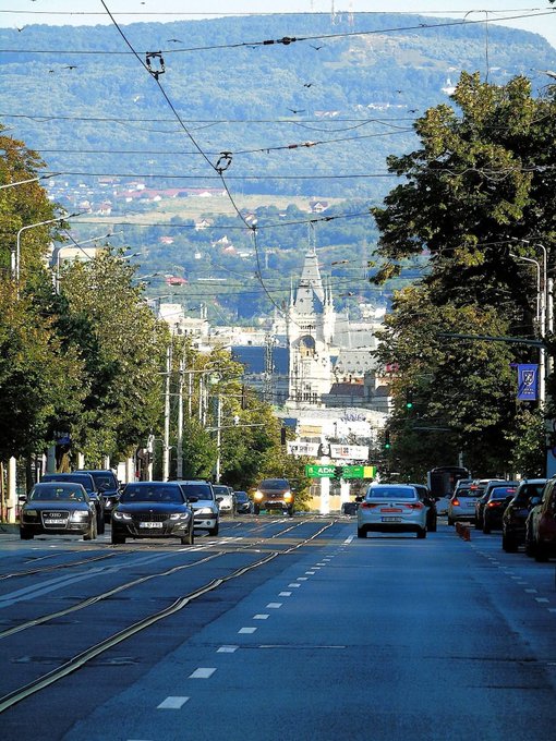 A scenic city street view looking toward an ornate cathedral with mountains in the background, featuring tram tracks, traffic, and overhead wires.