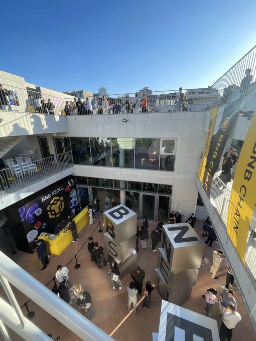 An elevated view of an outdoor event space called 'BNB CHAIN' with crowds of people gathered around promotional displays, including large 'BNB' cubes and a digital screen.