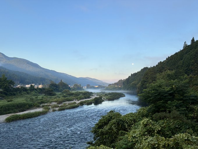 A scenic landscape photograph shows a river flowing between green, tree-covered hills and mountains under a clear sky with light mist on the water.