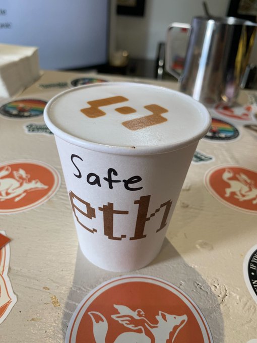 A white paper cup labeled 'Safe ETH' in pixel/blockchain-style font sits on a table decorated with stickers, including an orange winged boar logo.