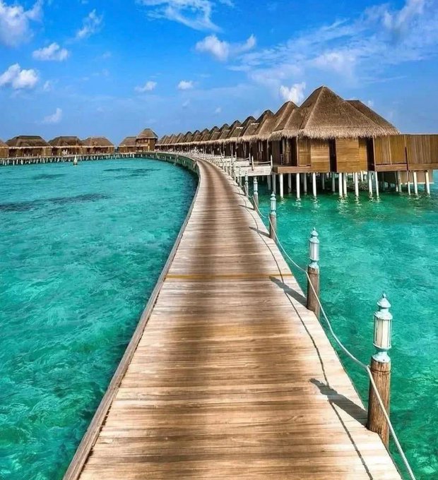 A wooden boardwalk extends across crystal-clear turquoise waters, leading to a row of thatched-roof overwater bungalows under a bright blue sky, resembling a tropical Maldives resort.