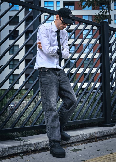 Street style photo of a young man in a white button-up shirt, black tie, dark grey distressed jeans, black sneakers, and a black cap, standing with crossed arms against a metal fence with apartment buildings in the background.