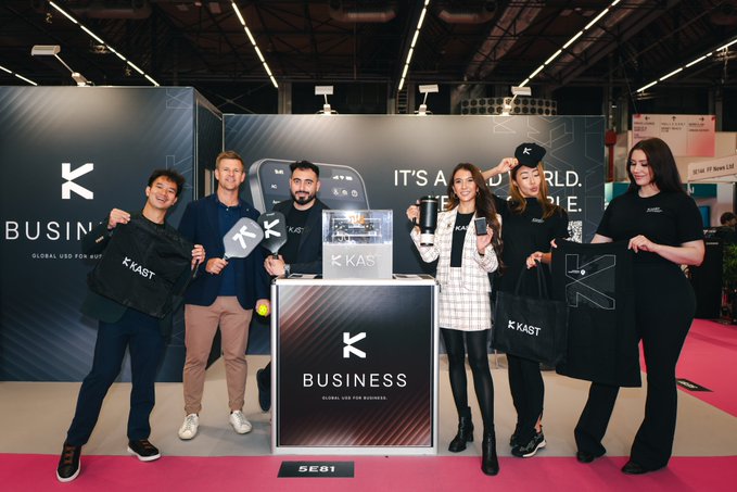 The image features a group of five people standing next to each other, all holding bags with "Business" written on them. They are posing together at an event or convention, possibly promoting a business idea or product. The background includes a booth for a company named "KAST XYZ", which is related to the theme of their gathering. The image also contains text that reads: "Global business never sleeps. Your money shouldn' t either. Traditional banking creates friction. @KAST XYZ removes it. 24/7 access. Borderless payments. Built for modern businesses." This suggests a connection between the group's presence and the company they are promoting, as well as their shared interest in the concept of "Business" being discussed at this event or convention.
