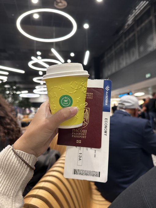 Photo of a hand holding a yellow coffee cup and a burgundy Romanian passport along with an Aeroflot boarding pass, taken in what appears to be an airport terminal.