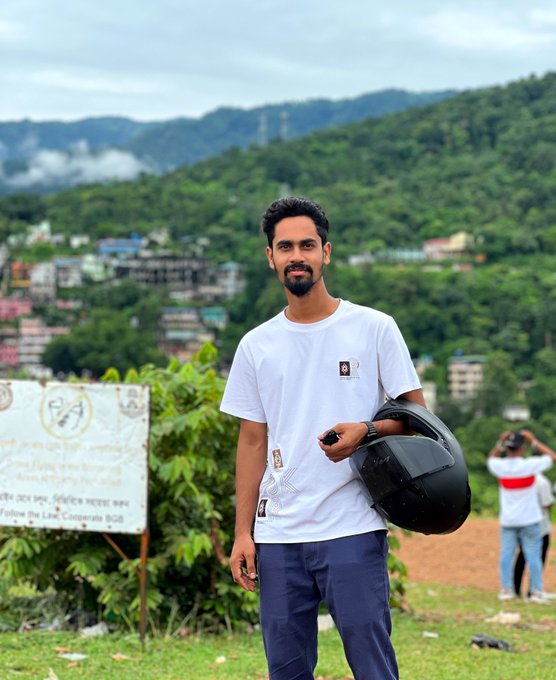 A young man with a beard stands outdoors in a white t-shirt, holding a black motorcycle helmet, with a hilly landscape and buildings visible in the background.