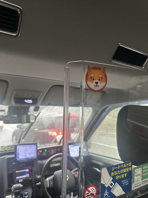 A view from inside a Japanese taxi with a clear protective partition displaying a Shiba Inu dog sticker on it.