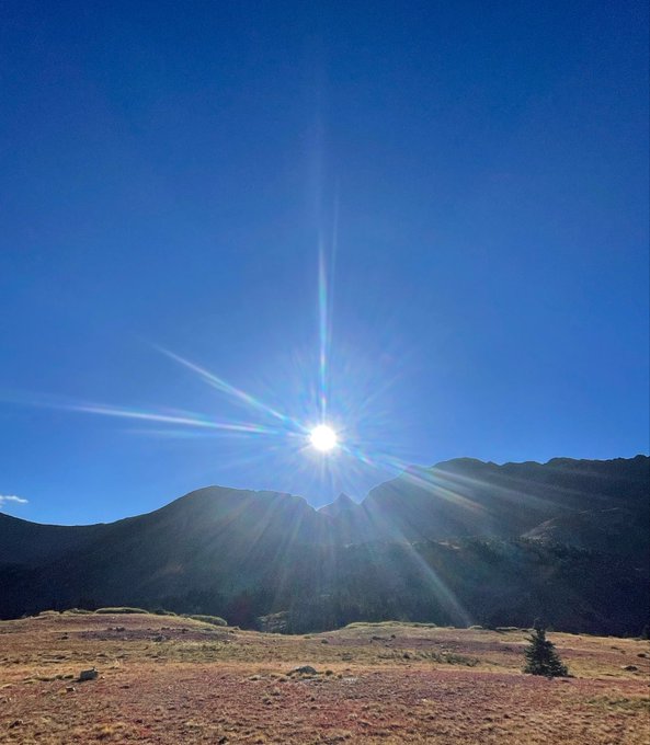 A scenic landscape photograph showing bright sun rays bursting over a mountain ridge under a clear blue sky, with reddish-brown grassy terrain in the foreground.