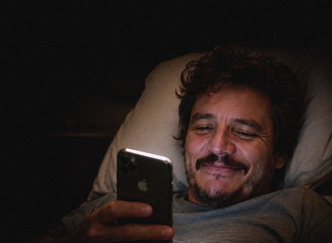 A man lies in bed smiling warmly at his smartphone in the dark, the Apple logo visible on its back. The scene captures a quiet, contented moment that mirrors the post's theme of simple, genuine happiness found in seeing friends succeed through ordinary life accomplishments rather than crypto hype.