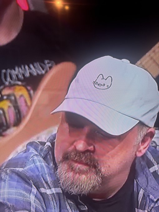 A bearded man in a blue plaid shirt is shown with a slight smirk, wearing a light blue baseball cap featuring the recognizable Cool Cats NFT logo on the front. The post references Redban appearing on the Kill Tony podcast wearing this Cool Cats hat, suggesting a return of the show.