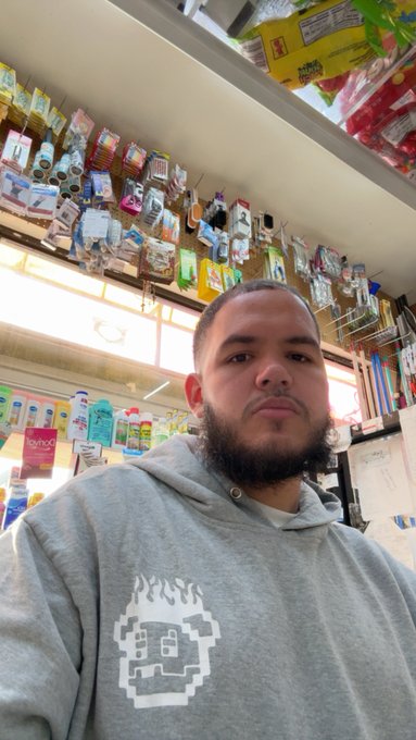 A bearded man wearing a gray hoodie with a pixelated graphic takes a selfie inside what appears to be a convenience store or small shop.