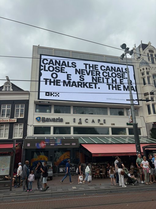 A large OKX billboard in Amsterdam displays the bold message "CANALS CLOSE. NEVER CLOSE. TOES NEITHE[R]. THE MARKET. THE MA[RKET]." above a Bavaria-branded building in a busy tourist area, with crypto exchange OKX reinforcing the post's observation that crypto advertising is widespread across the city.