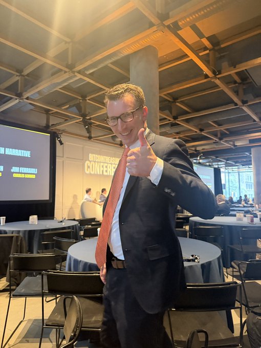 A man in a suit and orange tie gives a thumbs-up at what appears to be a storytelling conference, with a screen in the background showing "IN NARRATIVE" and "JIM FERRISOLI."