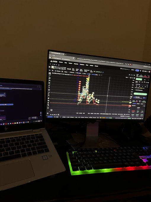 Laptop and RGB mechanical keyboard setup on a dark desk, displaying a TradingView-style candlestick chart on the monitor and a chat window on the laptop screen.