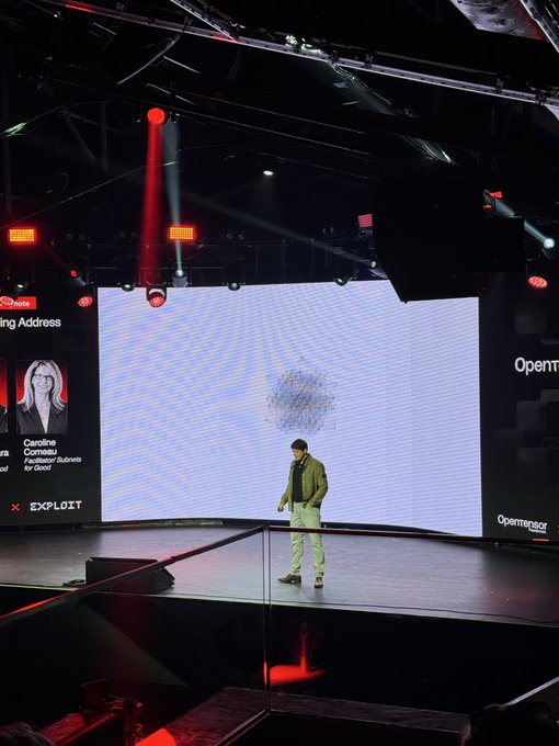 A man stands on stage at a tech conference, with a large screen displaying the "Opening Address" by Caroline Comeau, Founder/Robonist for Good, sponsored by Deutsche Bahn and EXPLOIT, alongside partial text reading "Openn" and "Opensea." The minimalist, futuristic setting with its dramatic lighting aligns with the post's mention of $TAO and the futuristic tech theme of @const_reborn's R.E.B.O.R.N project.