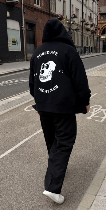 A person walking down a city street wearing a black "Bored Ape Yacht Club" hoodie with the iconic skull logo and "NYC" lettering, paired with black pants and white sneakers.