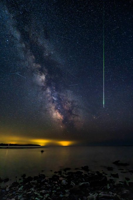 A night sky photograph featuring the Milky Way galaxy, stars, and a green meteor streaking over a calm body of water with rocky shoreline.